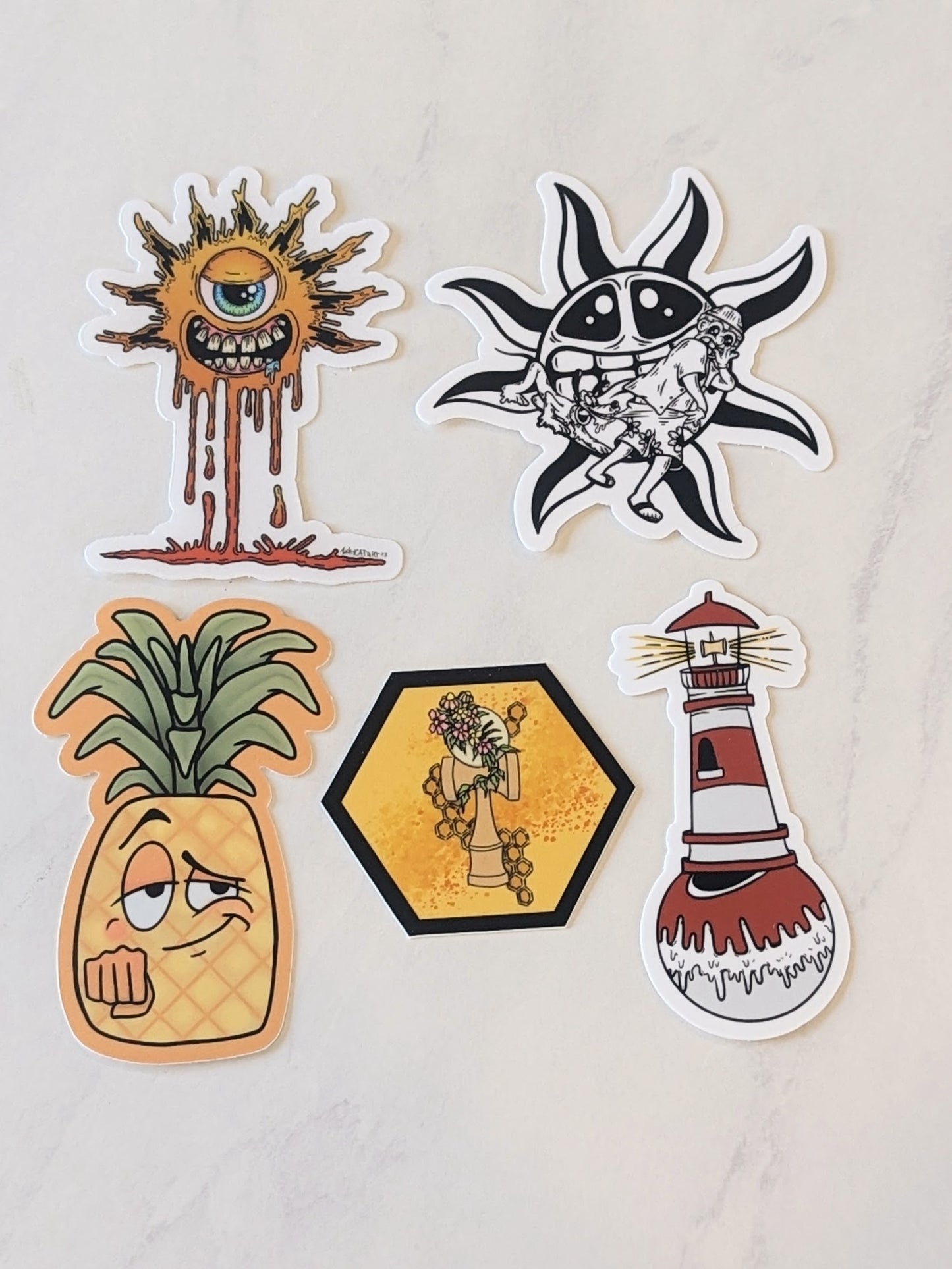 A collection of summer themed stickers by artists Kellie.love, Inkycat art, and Ghosttown ATL. The stickers include a cycloptic smiling orange drippy sun, A Black and white cartoon Sun grimmacing behind a dog pulling the surf shorts of a person gasping, a pineapple waiting to fist bump, a hexagon floral kendama illustration, and a lighthouse on a drippy tama (in a kendama lighthouse trick). 