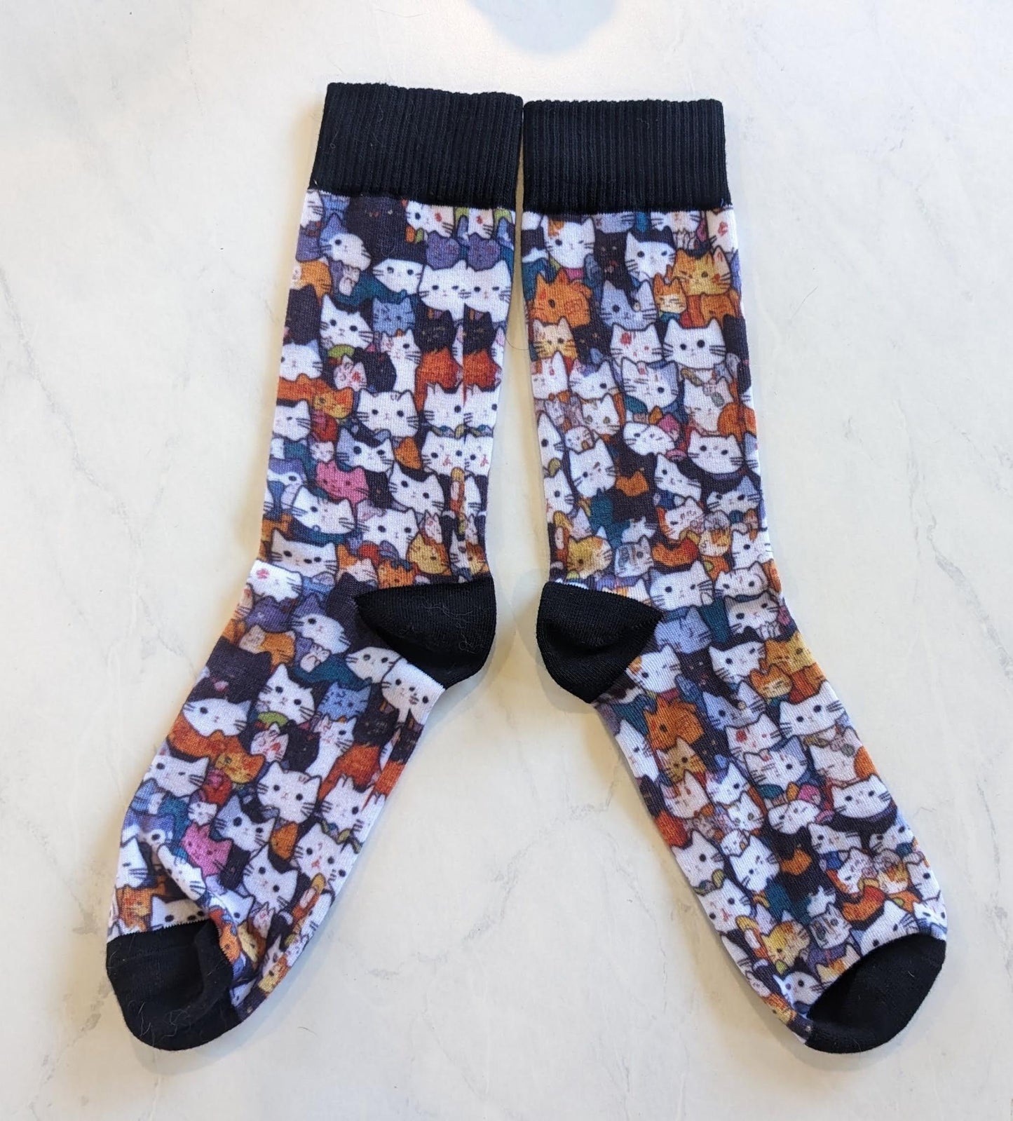A pair of socks with black toes, heels, and top. The socks otherwise are patterned with AI-generated cat heads that look like cute illustrations of a bunch of different cats. The cats come in a variety of colors, black, white, orange calico, grey, white and grey, and a few that stand out as colored differently (like pink or having a green spot).