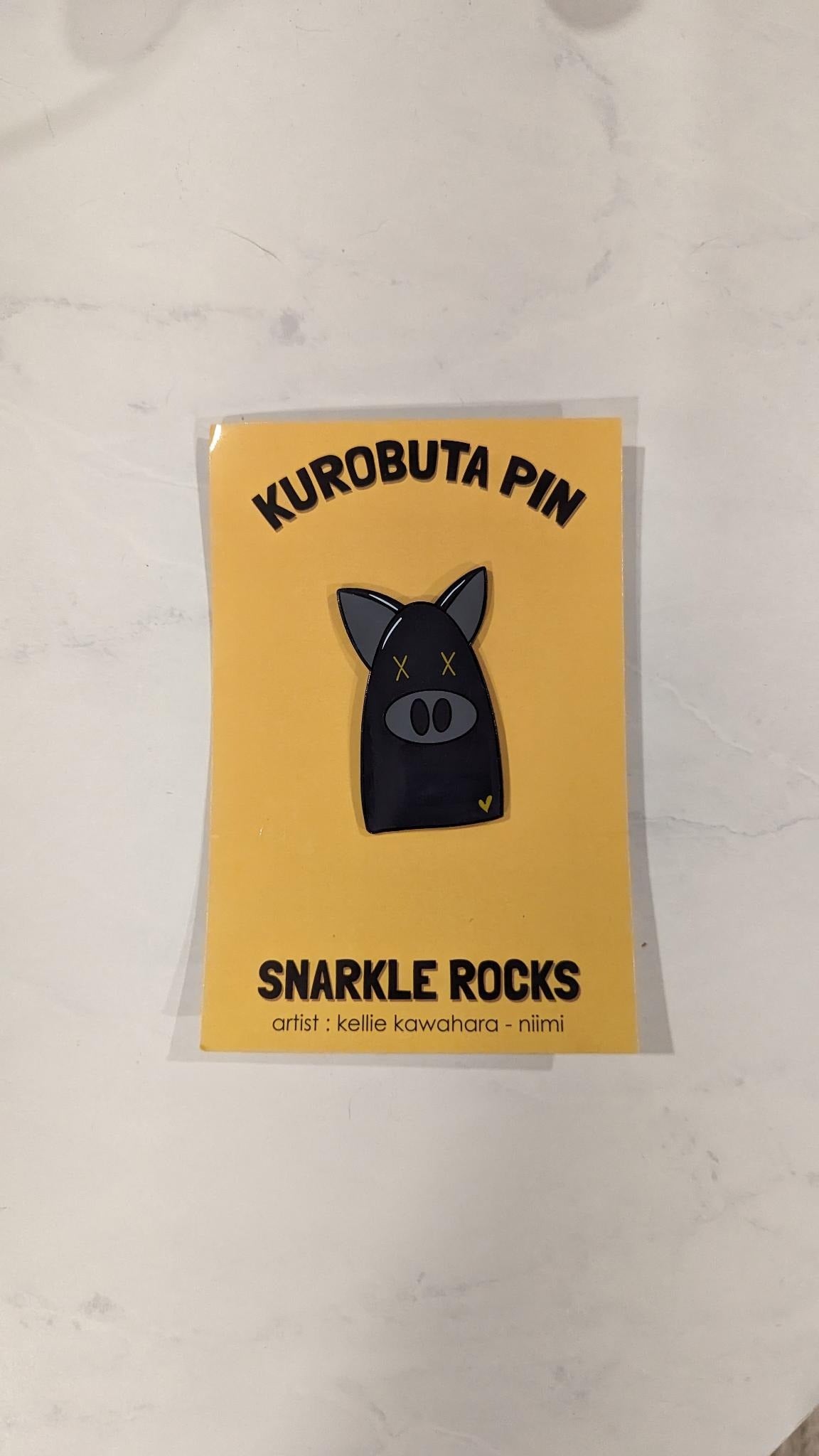 An enamel pin of a black pig with yellow X's for eyes and a yellow heart in the lower right corner.  The pin sits against a yellow cardstock backing with the text "Kurobuta pin" "Snarkle Rocks" "artist: kellie kawahara-niimi"