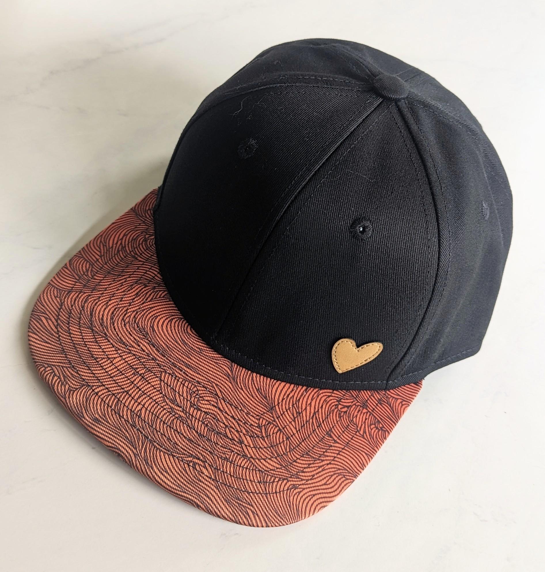 A black hat with a light tan leather heart sewed onto the front. The heart is about 1" tall and positioned in the lower right hand side if you are facing the front of the hat. The brim of the hat is a slight gradient of orange hues filled with thin, intricate organic black flowing wavy lines.