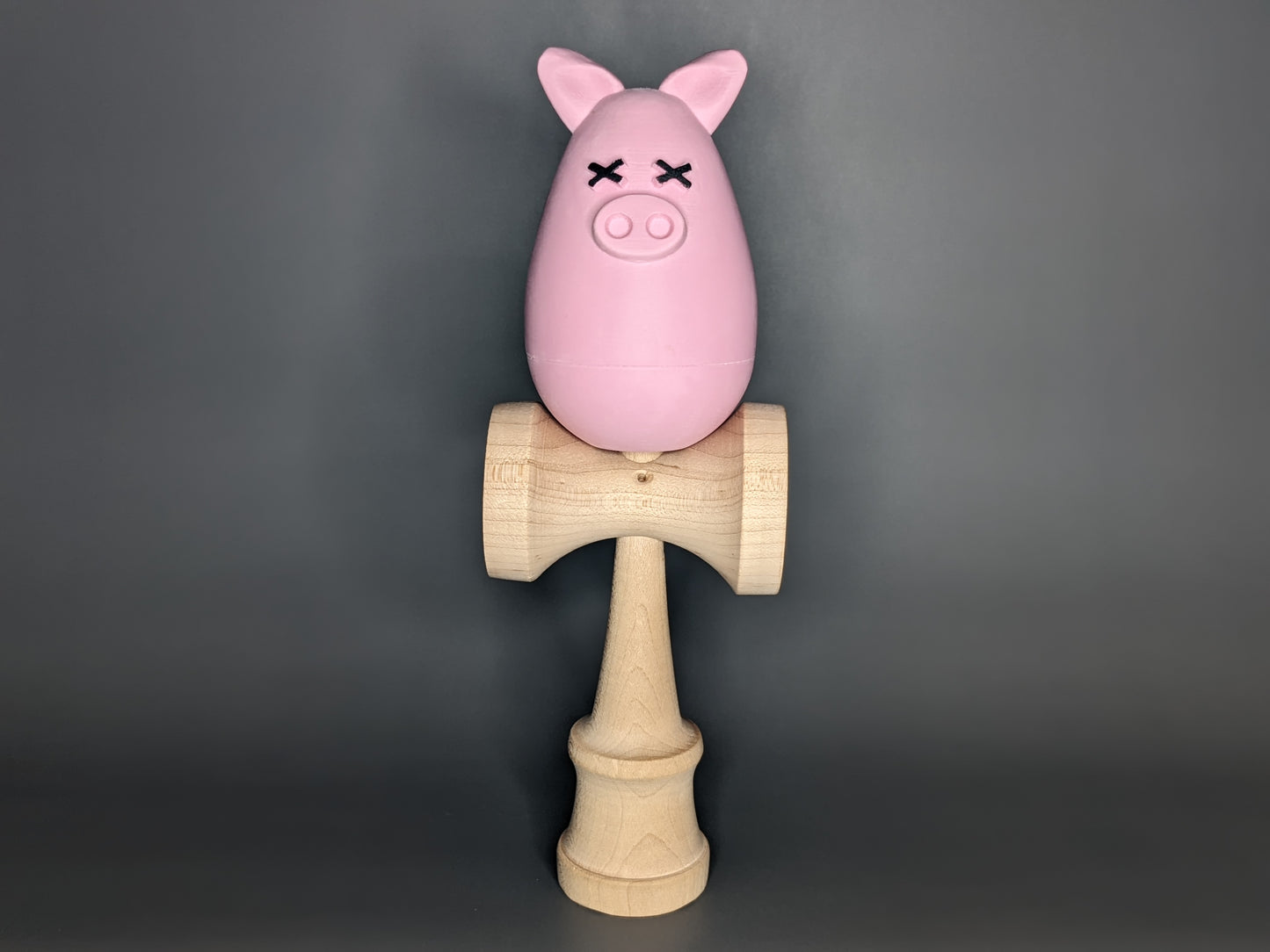 Pink 3D Printed Tama in an egg-like shape with ears, a pig snout and black x's for eyes (Kalua the Peeg). The 3d printed tama sits on a wooden Ken for a Kendama.